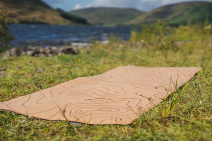 ROOTD Elite Terra Design. Non-toxic eco cork exercise mat topographical topo map design. Laying on grass next to a lake and mountains in the background. 