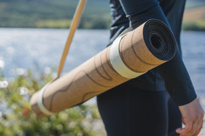 ROOTD elite mat Terra design. Non-toxic eco cork exercise mat over the shoulder with a white cotton carry strap and a topographical design mat. The background is around a lake and mountains. 