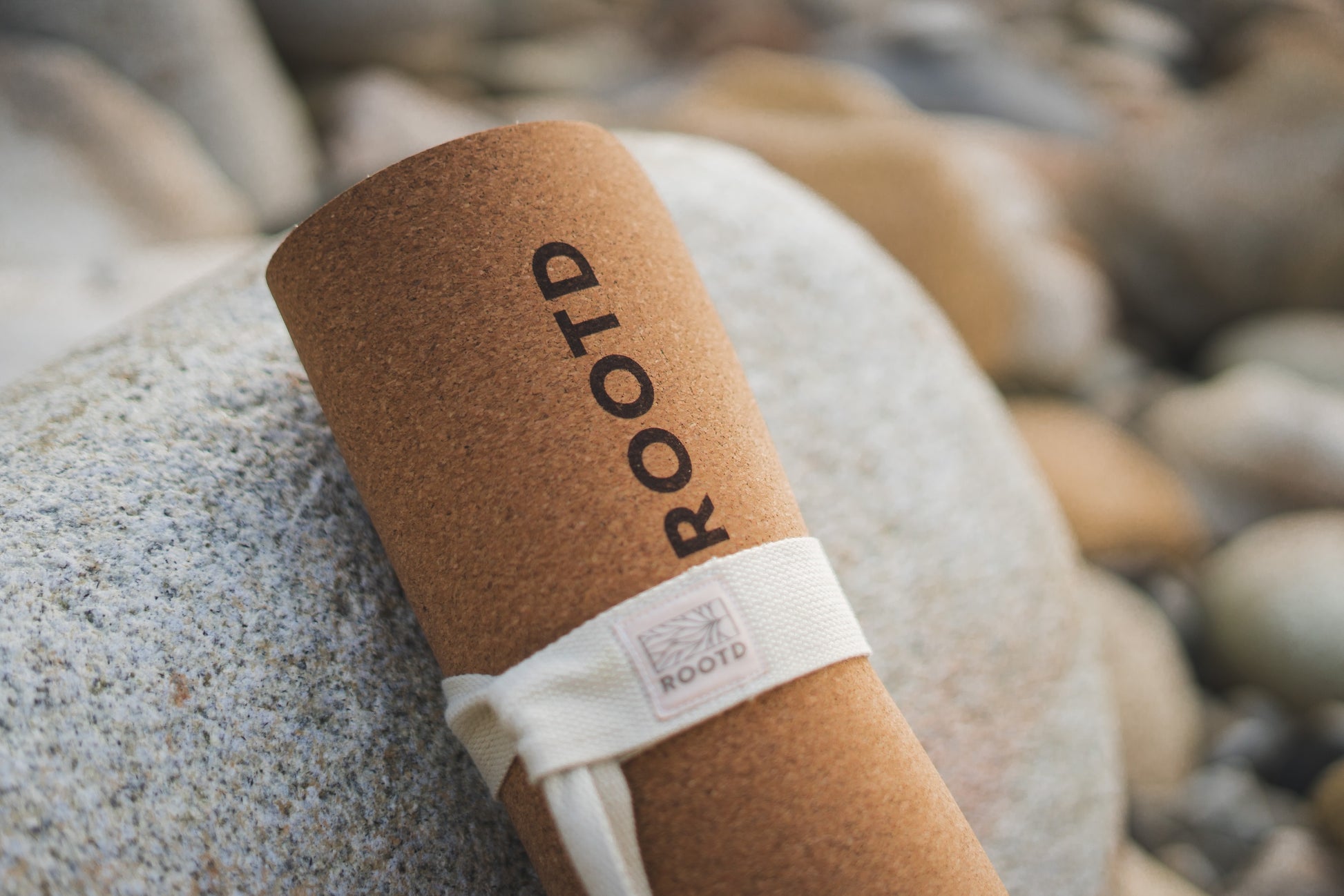 ROOTD Elite Bare Design. Non-toxic eco cork exercise mat with a plain minimal design. Rolled up with a white cotton carry strap. Propped up against a large stone at the beach.