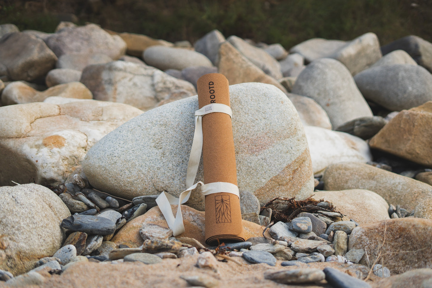 ROOTD Elite Bare Design. Non-toxic eco cork exercise mat with a plain minimal design. Rolled up with a white cotton carry strap. Propped up against a large stone at the beach.