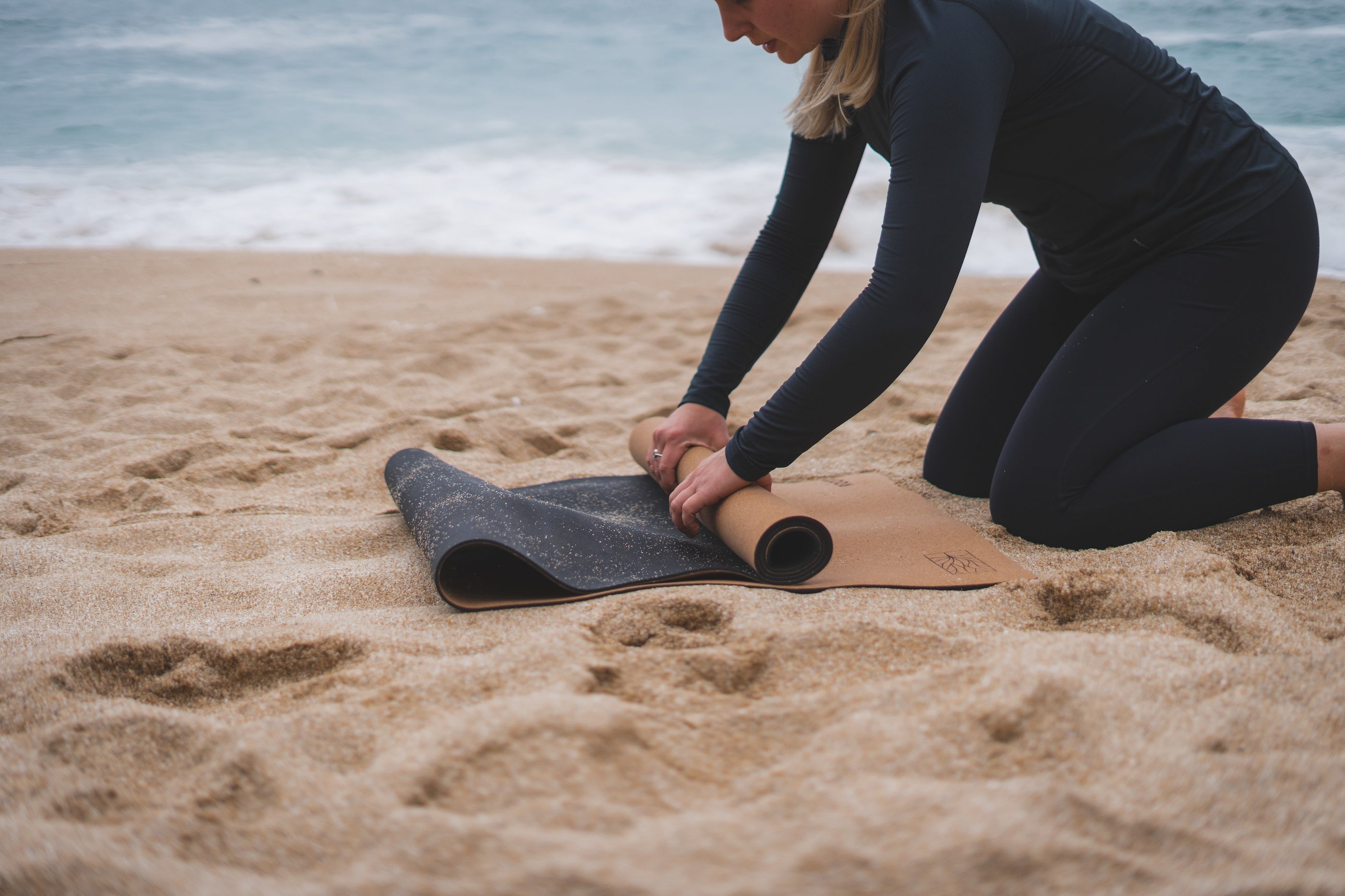 ROOTD Elite Bare Design. Non-toxic eco cork exercise mat being rolled up at the beach showing the rubber surface. 