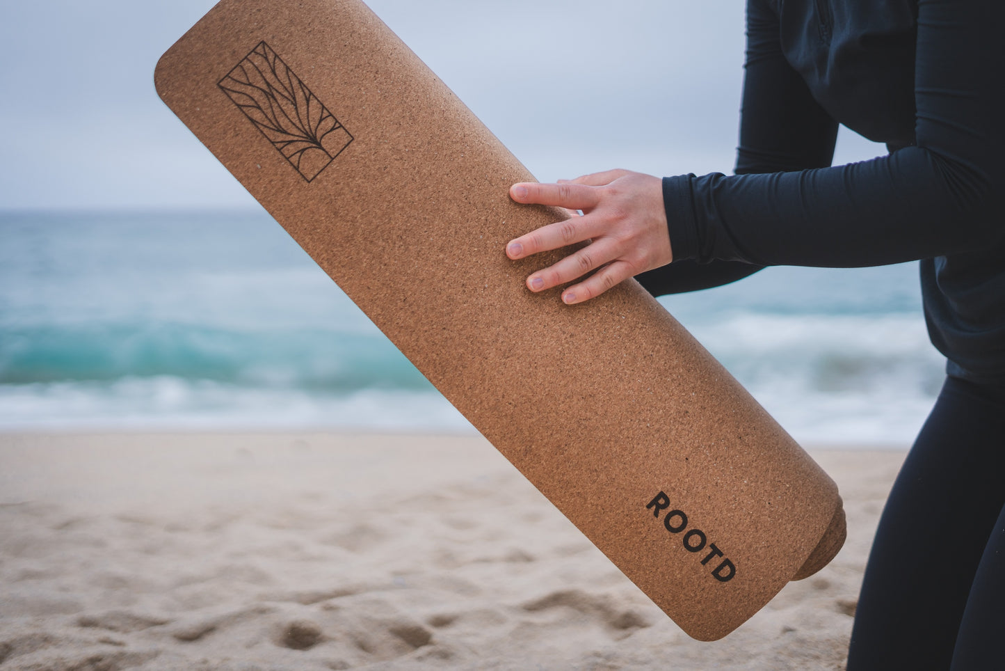 ROOTD Elite Bare Design. Non-toxic eco cork exercise mat with a plain minimal design. Woman rolling up the mat at the beach