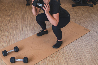 ROOTD Elite Align Design. Non-toxic eco cork exercise mat being used in a gym setting with dumbells and alignment lines. Woman doing squats.